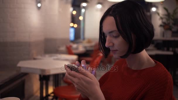 Chica joven caucásica usando teléfono inteligente sentado en la cafetería por la noche
 - Imágenes, Vídeo