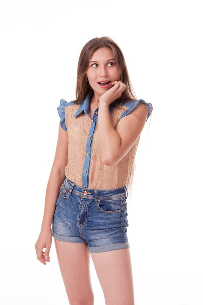 Beautiful young girl with long hair shows different emotions. Photographing on a white background.Photographing a girl with a beautiful figure in denim shorts and a blouse. having fun, sad, playing as an actor, posing for different poses.for advertis - Фото, изображение