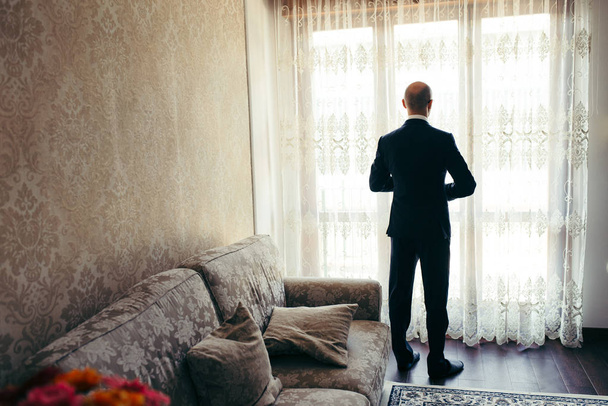 Full-length back view of the groom preparing for the wedding near the window. - Foto, Imagen