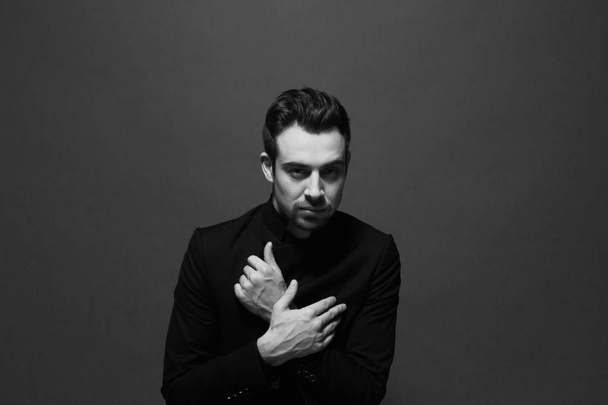 Black and white portrait of a young handsome man in all black suit, hands crossed on the chest, looking at the camera, against plain studio background. - Фото, зображення