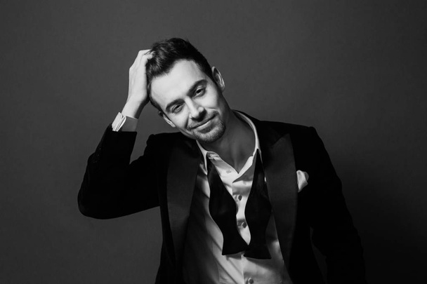 Retrato en blanco y negro de un joven guapo en traje, sonriendo y mirando a la cámara, pajarita y botones de camisa desabrochados
. - Foto, Imagen