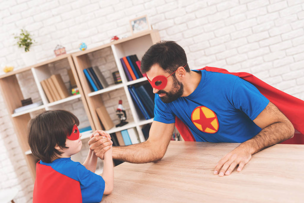 Father and son in suits of superheroes. They compete in armwrestling. They are measure their strength - Foto, afbeelding