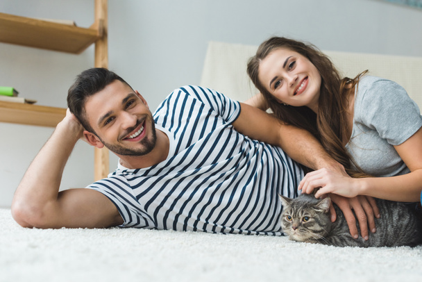 beautiful young couple lying on floor with adorable tabby cat - Foto, Imagen