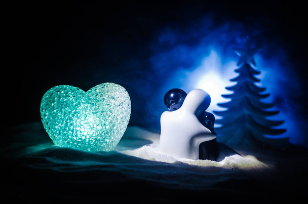 愛新年のコンセプトです。女の子と男の子の陶像は、白い雪と色心と暗い背景をぼかした写真のクリスマス ツリーの上に立って、お互い抱擁します。空の空間. - 写真・画像