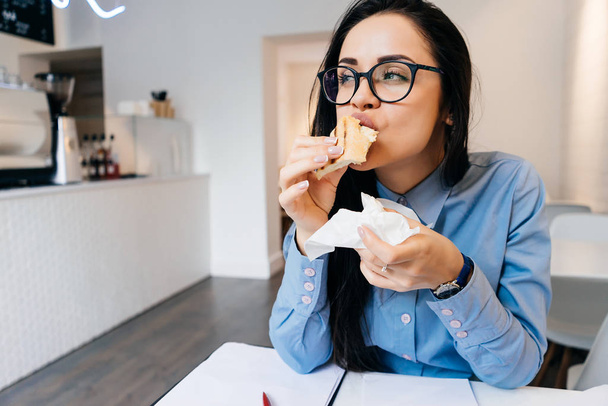 busy young girl with glasses eating breakfast in a cafe with a sandwich and coffee - Foto, imagen