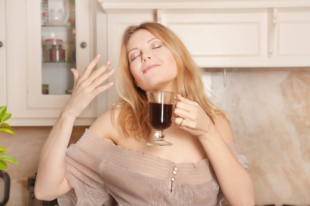 schöne Hausfrau mit geschlossenen Augen steht in ihrer weißen Küche und genießt morgens den starken Kaffeegeruch - Foto, Bild