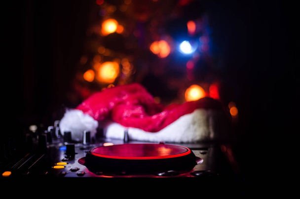 Dj Mixer with headphones on dark nightclub background with Christmas tree New Year Eve. Закройте вид на новогодние элементы или шприцы (Дед Мороз, Снеговик, Дог 2018, подарочная коробка) на столе у диджея. тонированные
 - Фото, изображение
