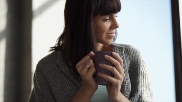 Młoda kobieta, picia kawy i patrząc przez okno - Materiał filmowy, wideo