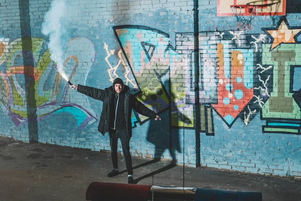 man holding smoke bomb and standing against wall with graffiti at night - Фото, зображення