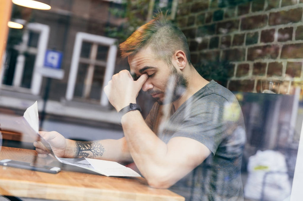 Mann, der mit Dokumenten in der Hand am Tisch sitzt und gestresst und müde wirkt. aus der Sicht hinter dem Fenster - Foto, Bild