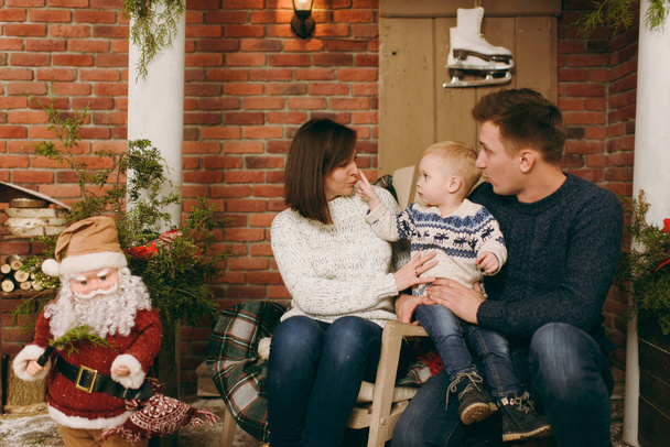Jovens pais felizes com um menino bonito criança vestida com suéter sentado na cadeira com o Papai Noel no quarto decorado Ano Novo em casa. Bom humor de Natal. Estilo de vida, família e férias 2018 conceito
 - Foto, Imagem
