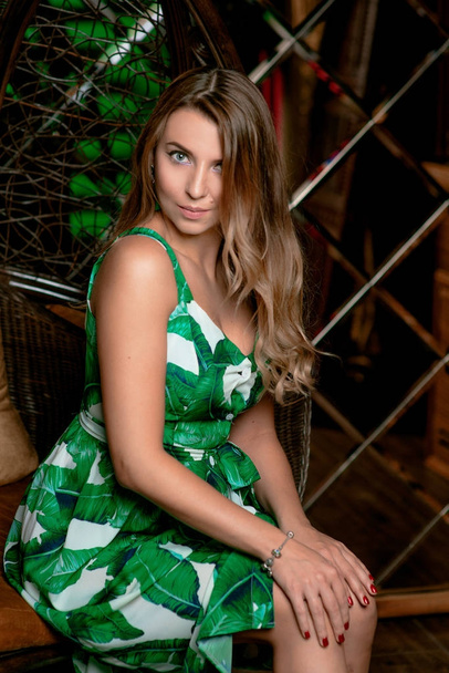 Portrait of stylish brown hair woman sitting in green tropical dress, green earrings, with makeup on bamboo armchair in the photostudio - Foto, afbeelding