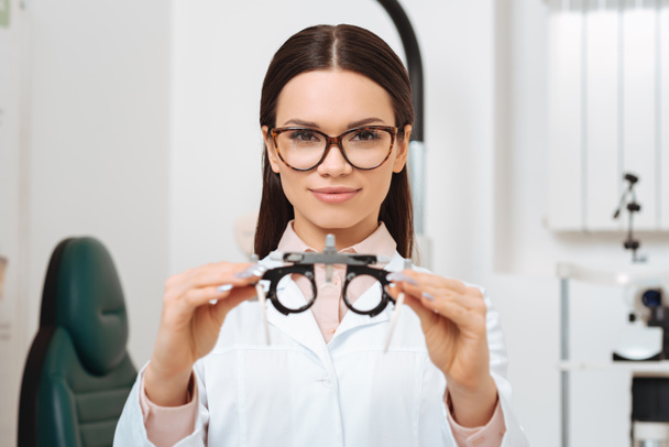 portrét mladé optometristy v bílém plášti, zobrazeno zkušební rám v rukou v klinice - Fotografie, Obrázek