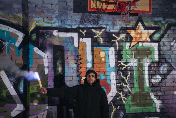 Человек держит дымовую бомбу и стоит у стены с граффити ночью
 - Фото, изображение