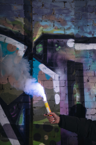 cropped view of hand with smoke bomb against wall with graffiti at night - Foto, Imagen