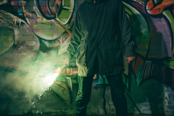 bijgesneden weergave van man houden rookbom en permanent tegen muur met graffiti nachts - Foto, afbeelding