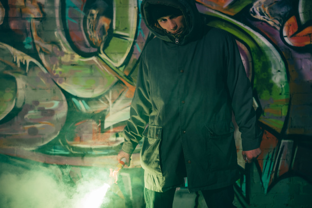 caucasian man holding smoke bomb and standing against wall with graffiti at night - Φωτογραφία, εικόνα