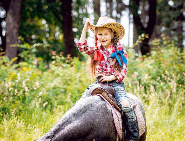 Little girl with braids in cowboy hat riding pony in park - Zdjęcie, obraz