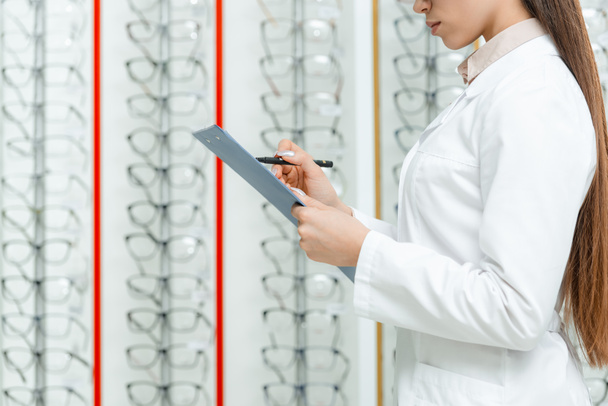 cropped shot of optometrist in white coat with notepad in hands standing in optics with eyeglasses on shelves - Foto, afbeelding