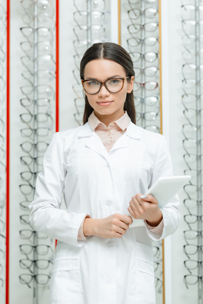 portrait of ophthalmologist with tablet in hands in optics with with eyeglasses on shelves - Photo, Image