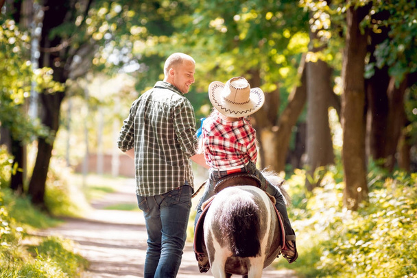 Little girl in cowboy hat riding pony with her father in park - Foto, afbeelding