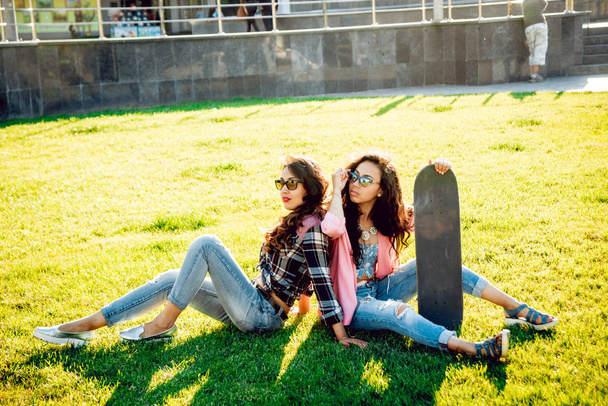 Two young mixed race girls in casual outfits with skateboard posing on green lawn - Фото, изображение
