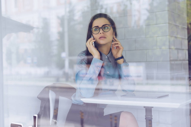 вдумчивая молодая девушка-фрилансер в очках работает в кафе, разговаривает по телефону о бизнесе
 - Фото, изображение