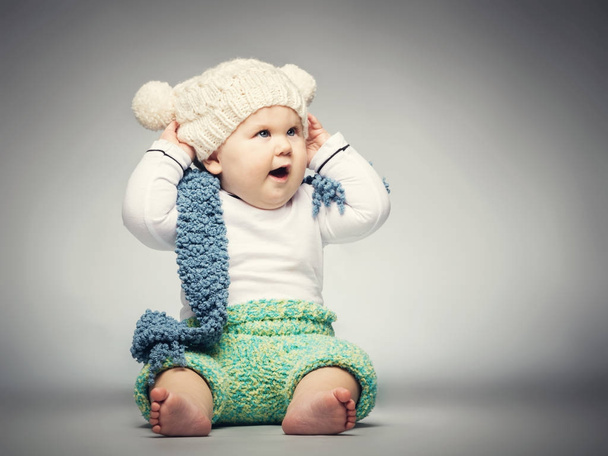 Kleine schattige jongen zittend op de grond, gekleed in wollen muts, sjaal en broek, op zoek gelukkig. - Foto, afbeelding