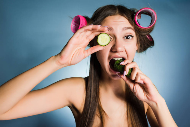 a funny young girl wants to look good, on the head of a curler, holds a cucumber to moisturize the skin on her face - Foto, imagen
