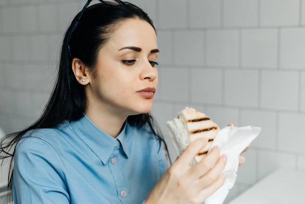 tired young girl student in blue shirt breakfasts sandwich in cafe after class - Фото, зображення