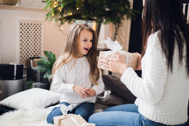 Merry Christmas and Happy Holidays. Cheerful mom and her cute daughter girl opening a Christmas present. Parent and little child having fun near Christmas tree indoors - Foto, afbeelding
