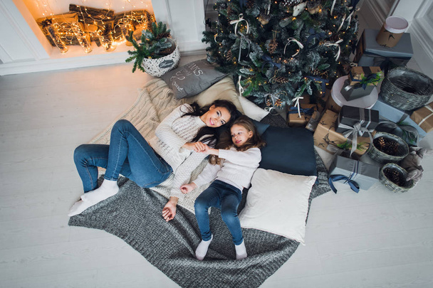 Forever best friends. Top view of happy mom and her daughter wearing jeans and white sweathers lying together on the blanket relaxing at home under christmas tree with gifts and fireplace nearby on a  - Zdjęcie, obraz