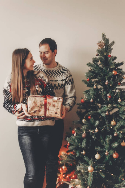 νεαρό ζευγάρι κρατώντας δώρο κουτί στέκεται κοντά στο χριστουγεννιάτικο δέντρο με φώτα - Φωτογραφία, εικόνα