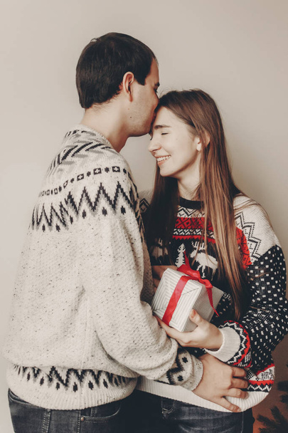 merry christmas and happy new year concept. stylish hipster couple in sweaters holding gift with red bow in room at christmas tree with lights and kissing. - Foto, Bild
