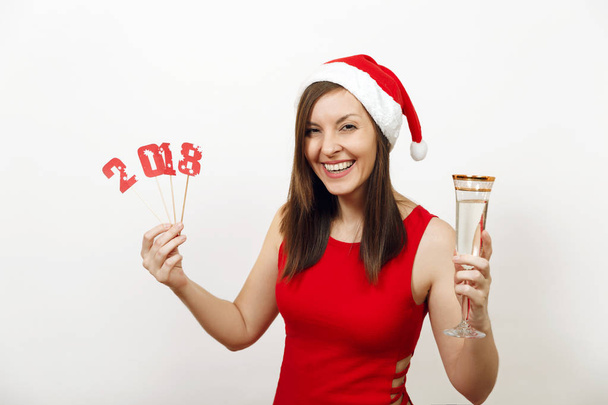 Kaunis valkoihoinen nuori onnellinen nainen viehättävä hymy punaisessa mekossa ja jouluhattu tilalla kortin numero 2018 ja lasi samppanjaa valkoisella taustalla. Joulupukin tyttö eristetty. Uusi vuosi loma
 - Valokuva, kuva