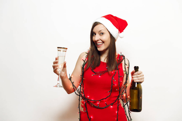 Beautiful charming young smiling happy woman in red dress, Christmas hat, garland lanterns standing with glass and bottle of champagne on white background. Santa girl isolated. New Year 2018 holiday - Photo, Image