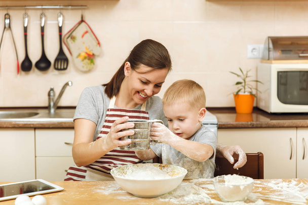 Little kid boy helps mother to cook Christmas ginger biscuit in light kitchen with tablet on the table. Happy family mom 30-35 years and child 2-3 in weekend morning at home. Relationship concept - Foto, afbeelding