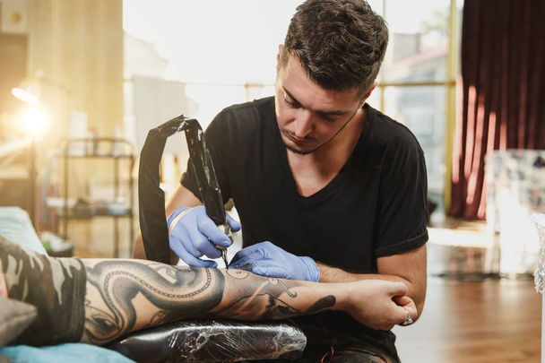 Szakmai tattooer művész csinál kép a kezét, az ember a gép fekete tinta a jar. A test tetoválás minták. A tetoválás művészet berendezés. Mester teszi tetovált világos stúdió. - Fotó, kép
