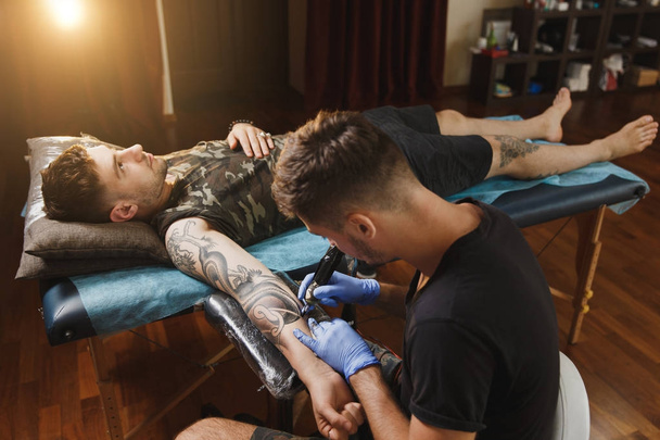 Artista profesional tatuador haciendo la imagen a mano del hombre por la máquina tinta negra de un frasco. Arte del tatuaje en el cuerpo. Equipo para hacer arte del tatuaje. Maestro hace tatuado en estudio de luz
. - Foto, imagen