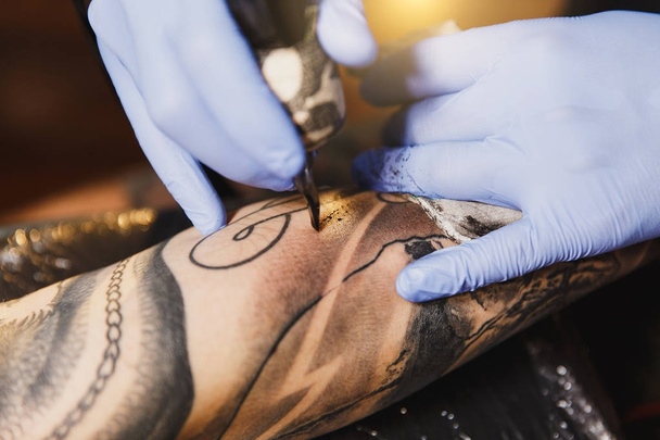 Közelről szakmai tattooer művész csinál kép a kezét, az ember a gép fekete tinta a jar. A test tetoválás minták. A tetoválás művészet berendezés. Mester teszi tetovált világos stúdió. - Fotó, kép