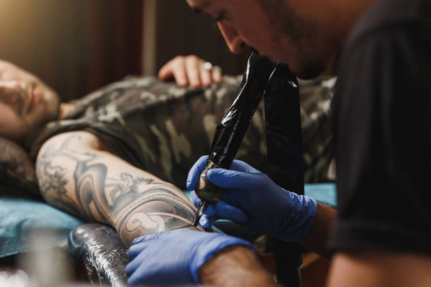 Close up van professionele tattooer kunstenaar foto aan de kant van de mens door machine zwarte inkt uit een potje doen. De kunst van de tatoeage op het lichaam. Apparatuur voor het maken van tattoo kunst. Master maakt getatoeëerd in lichte studio. - Foto, afbeelding