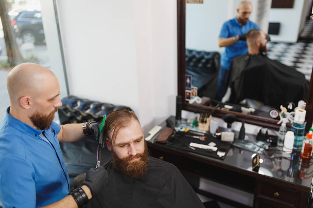 Male professional hairdresser serving client by scissors. Ginger handsome brutal stylish young man with thick big beard, short hair getting trendy haircut in black cape. Light white barber shop room. - Photo, Image