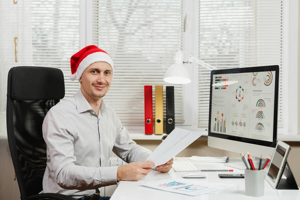 Handsome smiling business man in shirt and red Christmas hat sitting at the desk, working at computer with modern monitor, with documents in light office, looking at the camera on window background - Photo, Image
