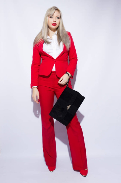 Portrait of a beautiful young blond woman with long hair in a red suit. She holds a big black clutch in her hand - Фото, зображення