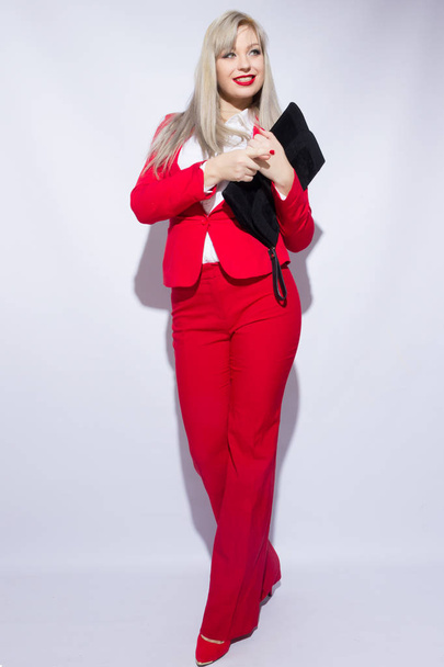Portrait of a beautiful young blond woman with long hair in a red suit. She holds a big black clutch in her hand - Fotó, kép