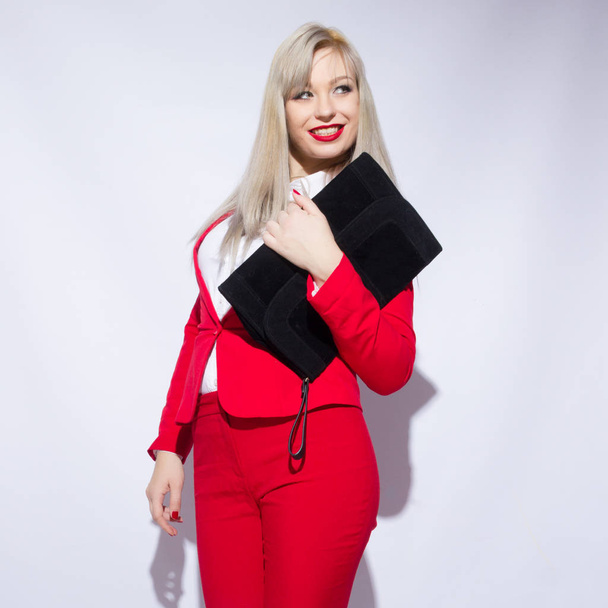 Portrait of a beautiful young blond woman with long hair in a red suit. She holds a big black clutch in her hand - Foto, Imagem