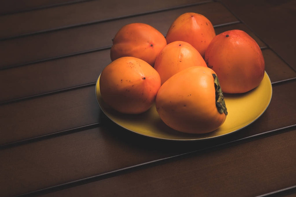 yellow plate with orange persimmons on brown table - Fotó, kép