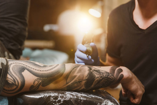 Close up of professional tattooer artist doing picture on hand of man by machine black ink from a jar. Tattoo art on body. Equipment for making tattoo art. Master makes tattooed in light studio. - Photo, Image