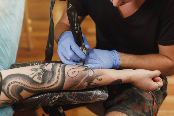 Крупным планом профессионального татуировщика, рисующего на руке человека черными чернилами из банки. Татуировка на теле. Оборудование для изготовления татуировок. Мастер делает татуировки в светлой студии
. - Фото, изображение