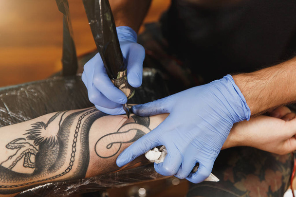Zbliżenie na profesjonalnych tattooer artysta robi zdjęcie na dłoni człowieka przez maszyny czarny tusz od słoika. Sztuka tatuaż na ciele. Sprzęt do tatuażu sztuki. Master sprawia tatuażem w studio światła. - Zdjęcie, obraz